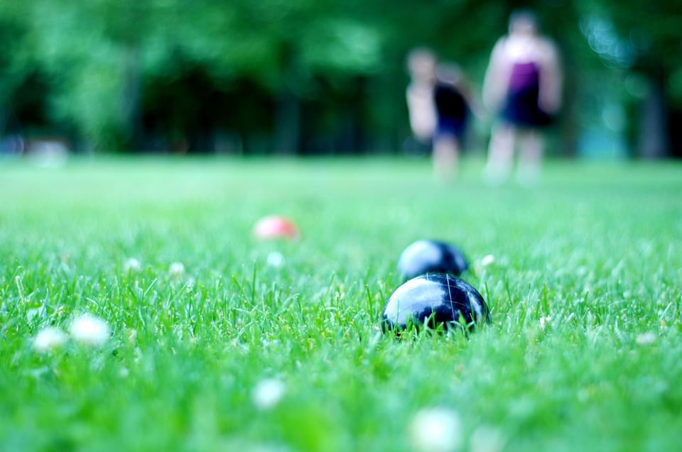 Black bocce balls resting on vibrant green grass with blurred family playing lawn games in background at Hotel X Toronto