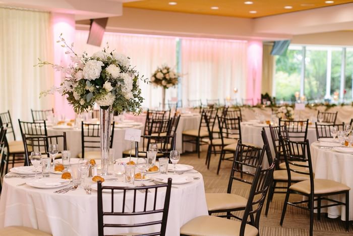 Dining area with tables and chairs, white tablecloths, floral centerpieces, and visible outdoor greenery
