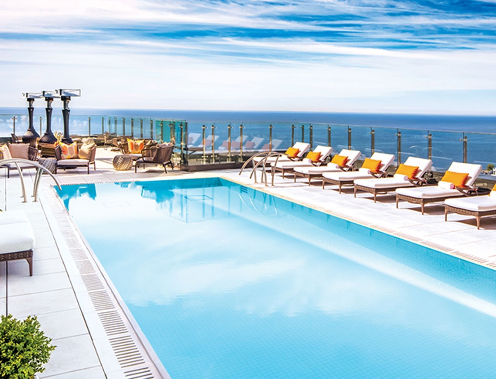 Rooftop pool lined by lounge chairs with orange cushions, under a glass railing overlooking the ocean at Hotel X Toronto
