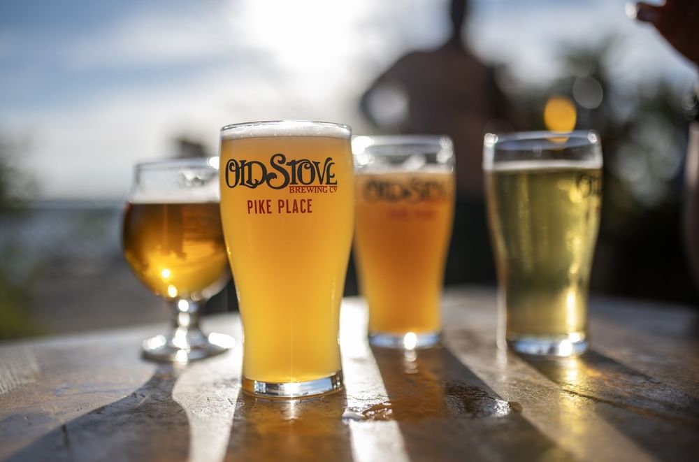 Four full beer glasses with a logo stand on a sunny wooden table at Warwick Seattle