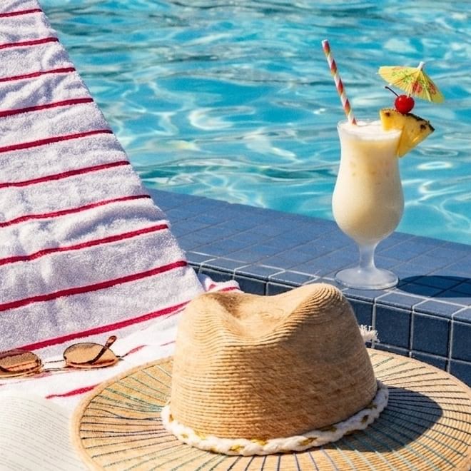 A still life by the pool with a drink, hat, sunglasses, and towel.