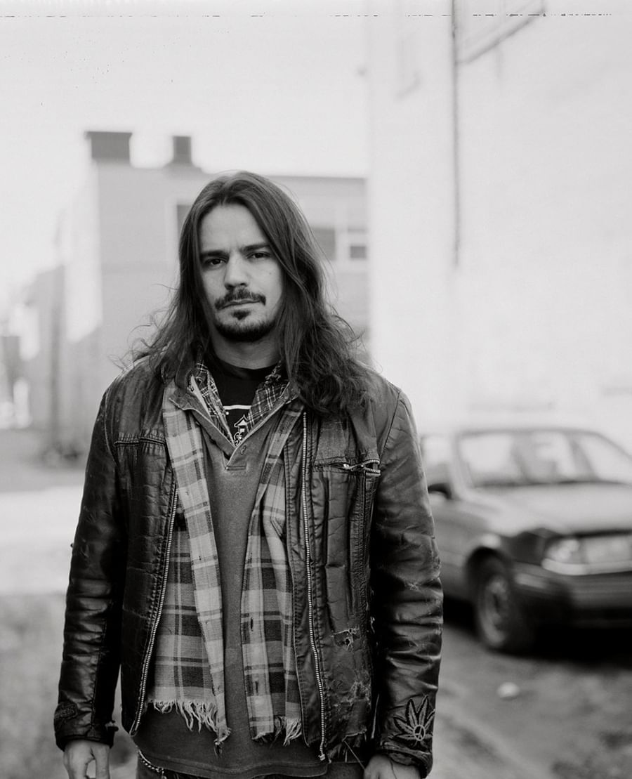 Black and white portrait of a man with long hair wearing a leather jacket and plaid scarf on an urban street at Warwick Le Crystal Montreal