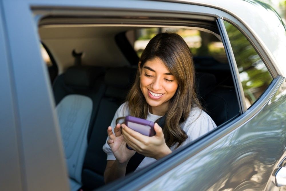 A view through the window of a car of a woman sitting in the backseat, smiling and looking at her phone.