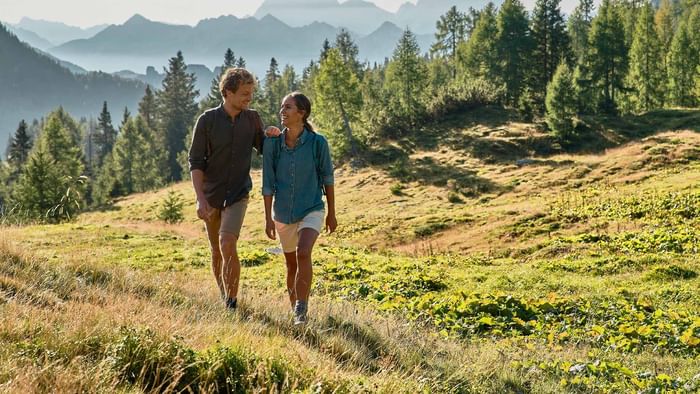 Couple walking on a grassy path through a forest with mountains in the background.