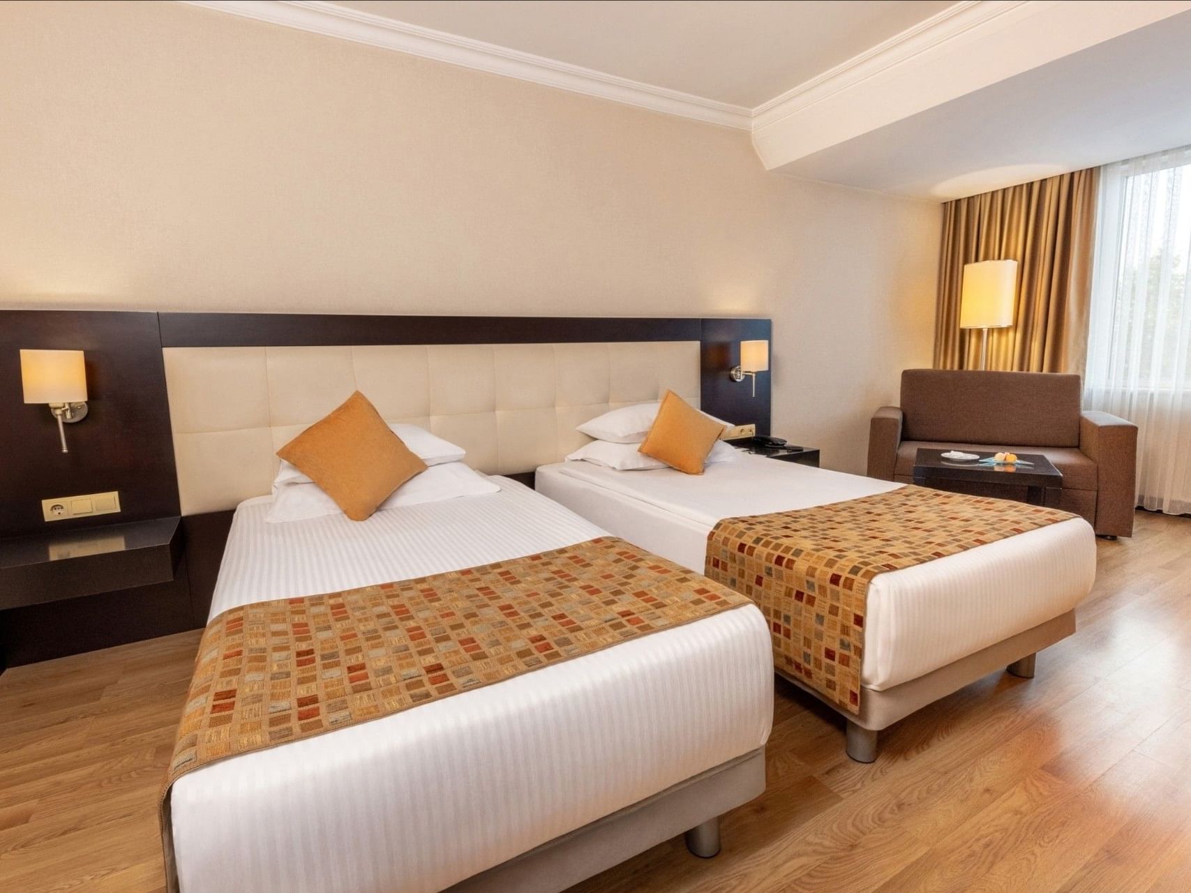 Zwei Doppelbetten in einem Hotelzimmer mit Holzboden, Couch und Fenster mit Vorhängen.