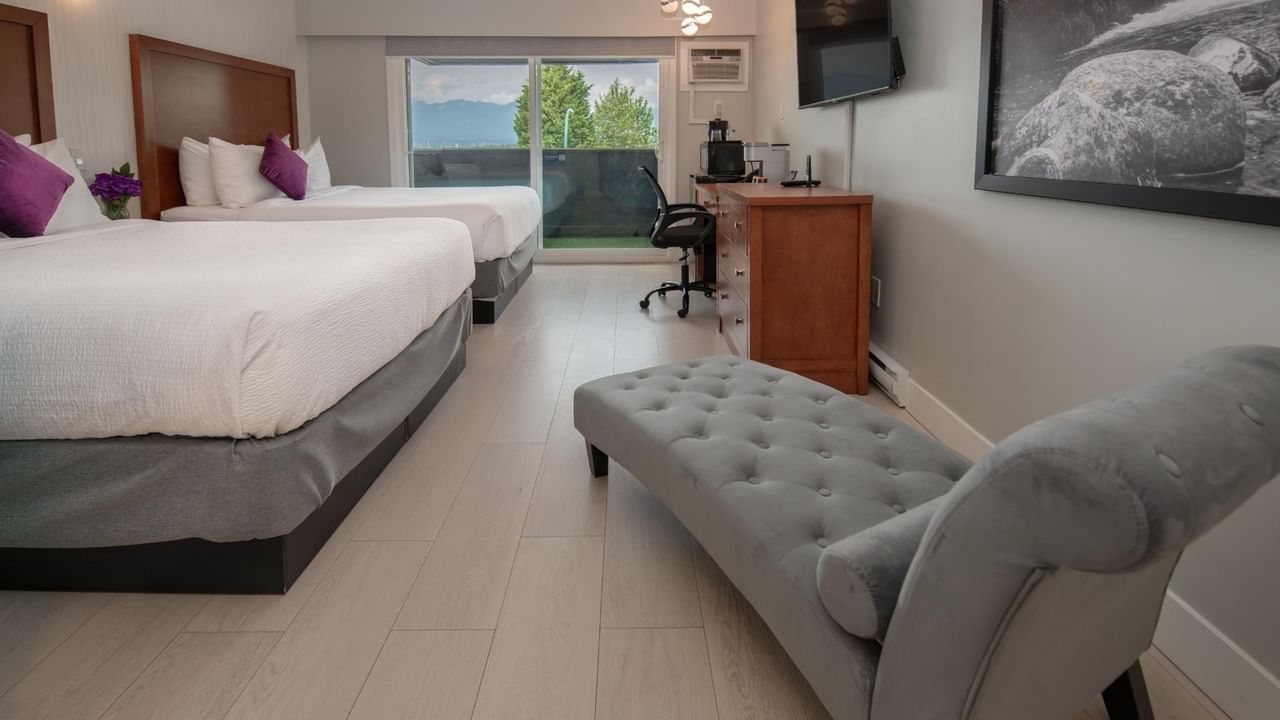 Two queen beds, a chair, a bench, a desk, a painting and a balcony in a hotel room.