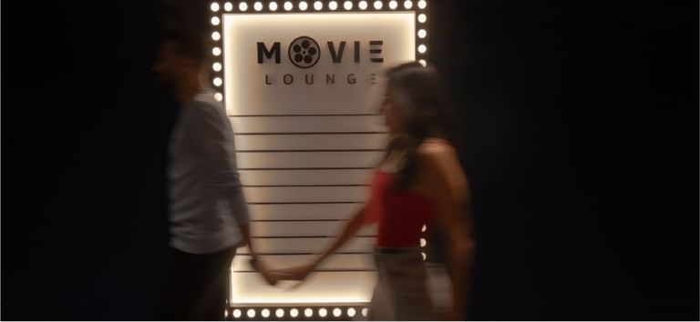 Couple holding hands walking past illuminated Movie Lounge sign creating cinematic vibes at DAMAC Maison Aykon City