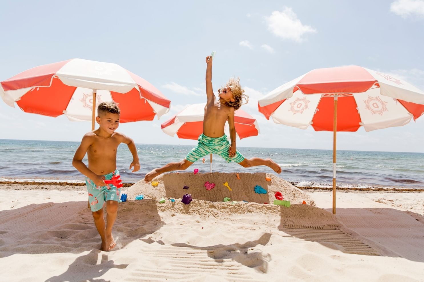 Two kids play joyfully on a sunny beach with sand sculptures and colorful umbrellas at The Diplomat Resort