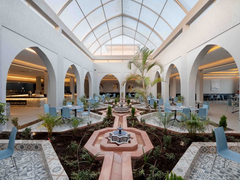 A modern indoor courtyard with arched windows, blue chairs, a central garden area, and a glass-domed ceiling.