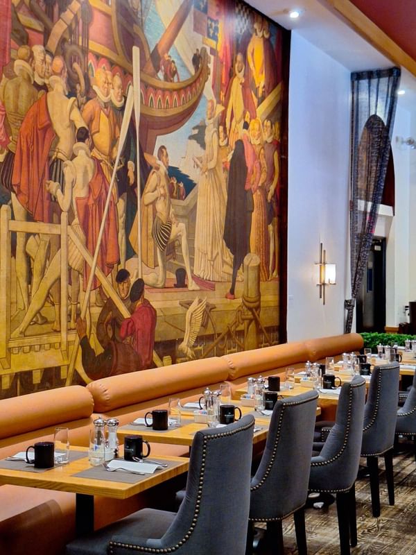 Long dining table with orange banquette seating positioned before large historical mural in Murals on 54 at Warwick New York
