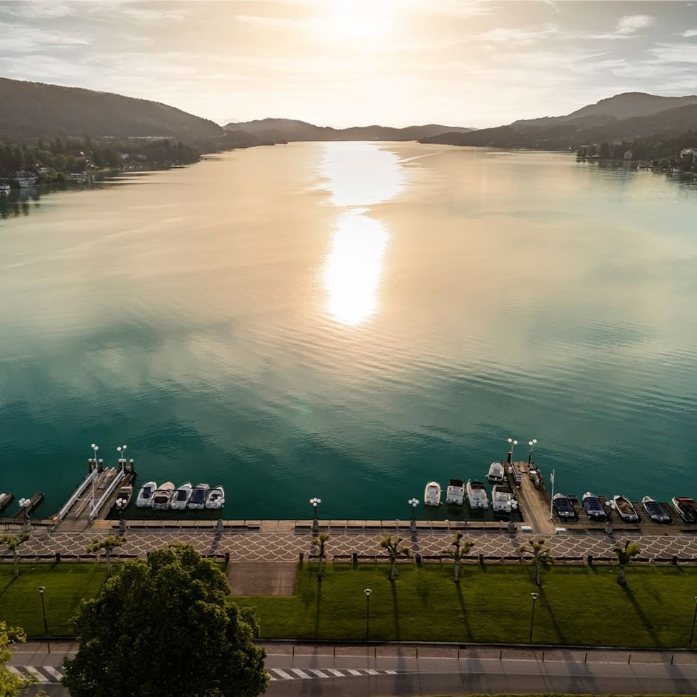Vista aerea di un lago al tramonto con barche ormeggiate e colline sullo sfondo.