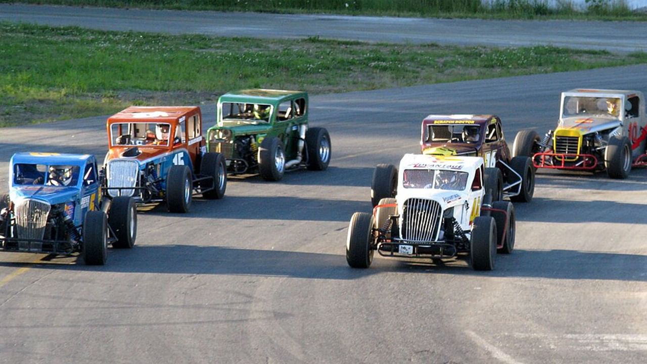 Six vintage race cars with drivers lined up on a racetrack with grass and trees in Penticton, B.C