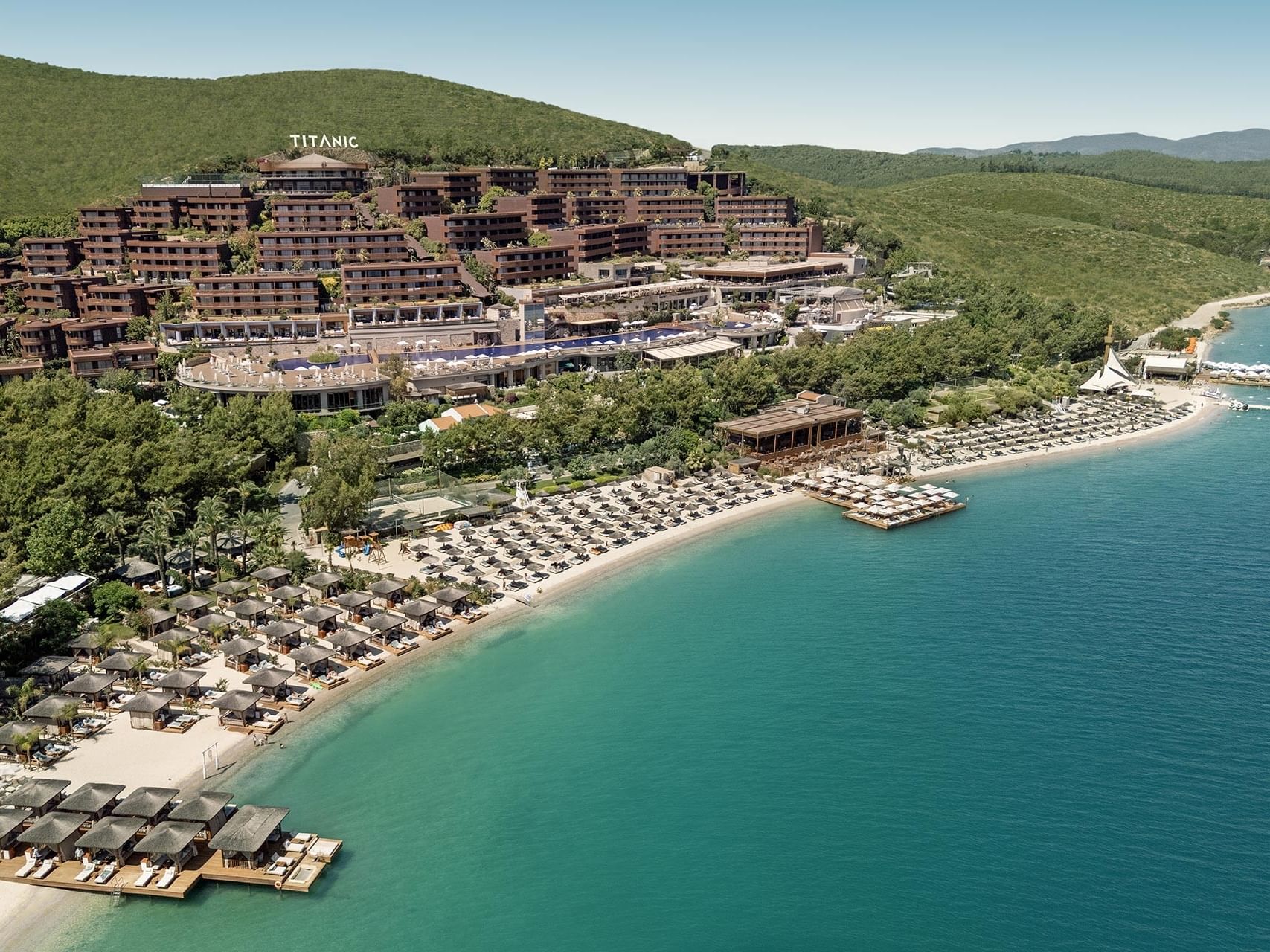 Aerial view of Titanic Luxury Collection Bodrum by the sea, featuring a beachfront and mountain scenery in the distance.
