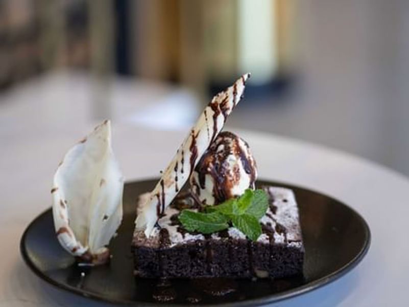 Dark brownie topped with ice cream and white chocolate by a mint leaf on a black dessert plate in KVNT at Warwick Al Jubail