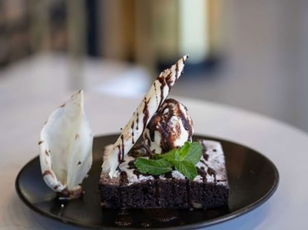 Dark brownie topped with ice cream and white chocolate by a mint leaf on a black dessert plate in KVNT at Warwick Al Jubail