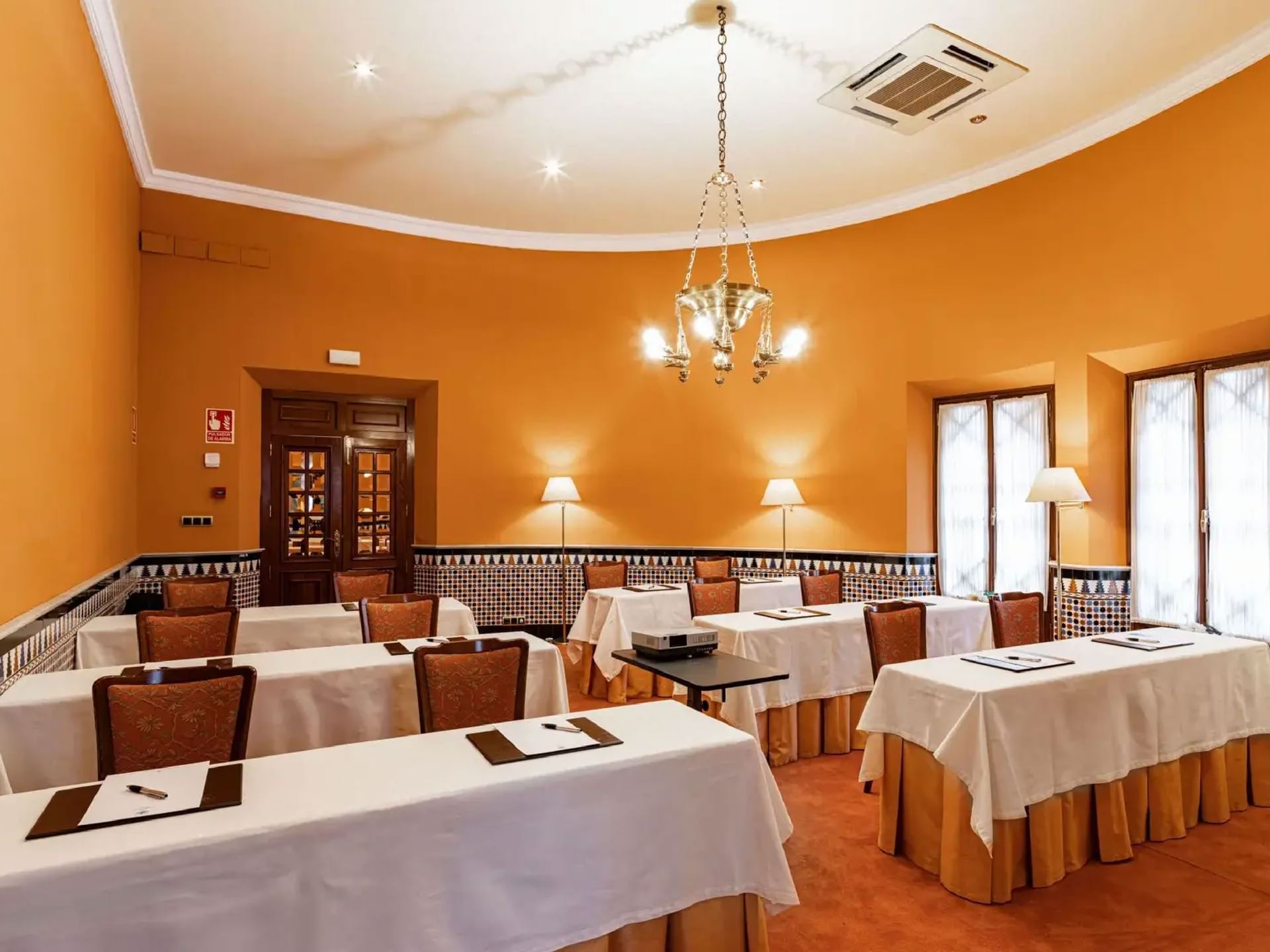 Brightly lit Sierra Nevada Room at Hotel Alhambra Palace, with orange walls, chairs, elegant chandeliers, and large windows