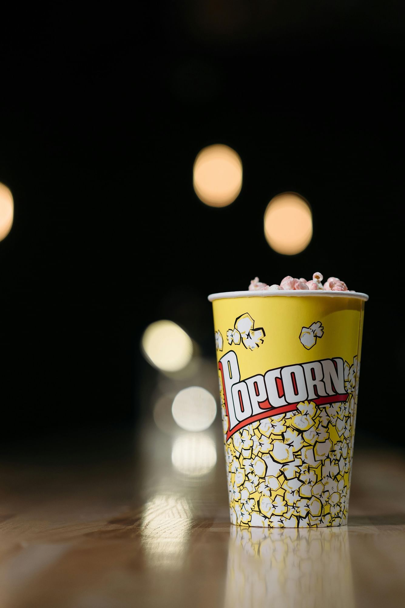A yellow bucket of popcorn sits on a shiny wooden surface with out of focus lights glowing in the background.