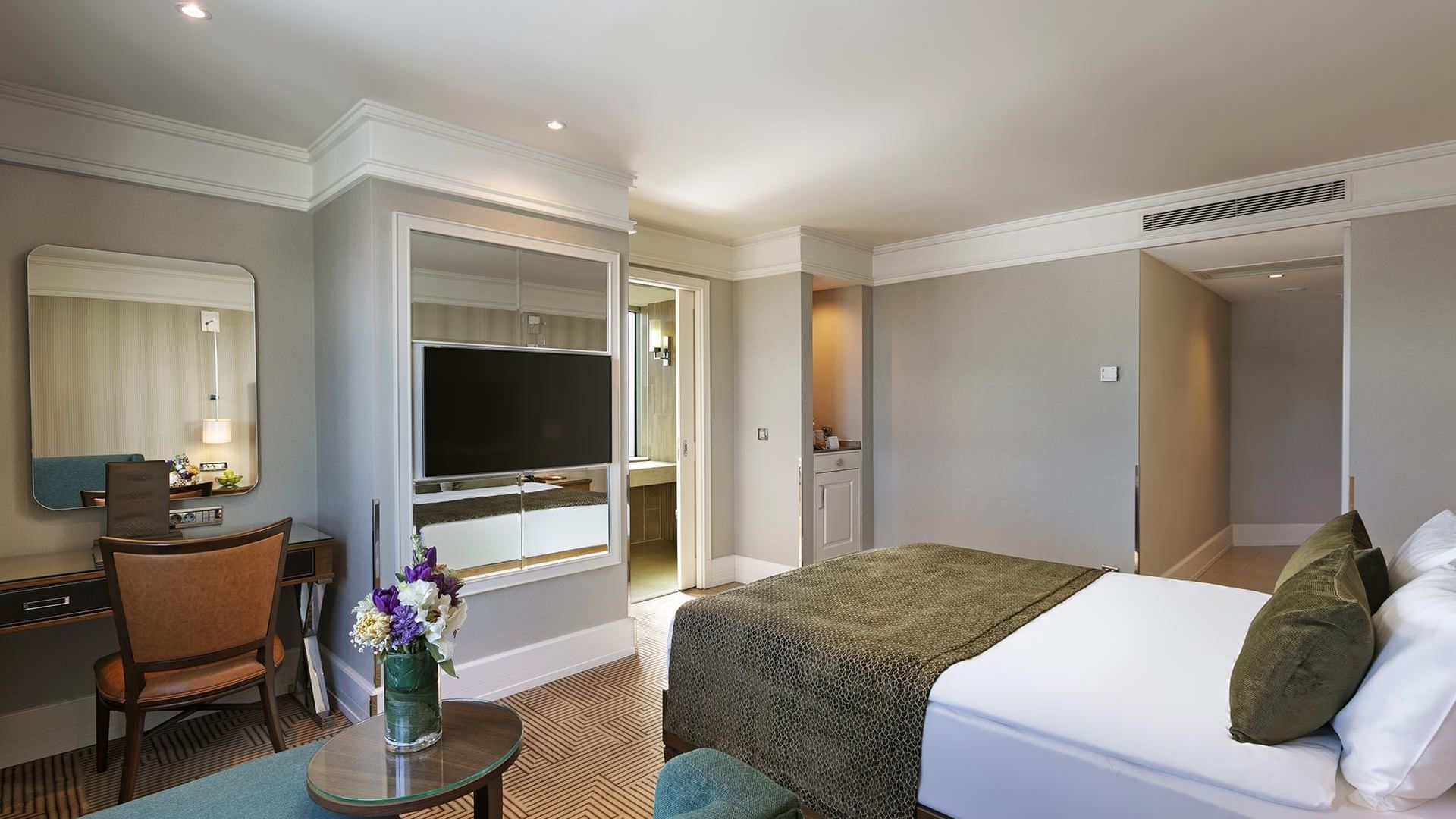 A hotel room with a bed, a chair, a desk, a mirror, a TV, and flowers on a table in Deluxe Room at Titanic Port Bakırköy