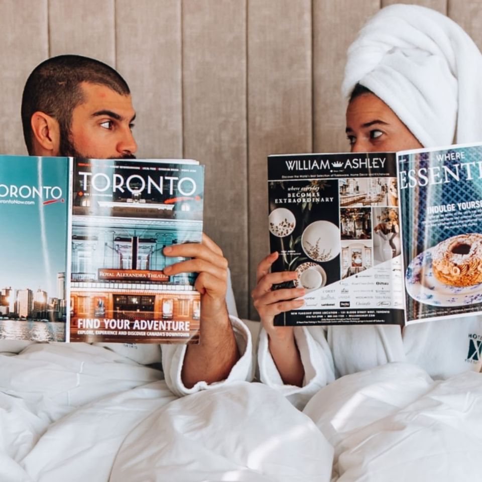 Couple in white robes reading Toronto magazines together by a padded headboard under soft lighting at Hotel X Toronto