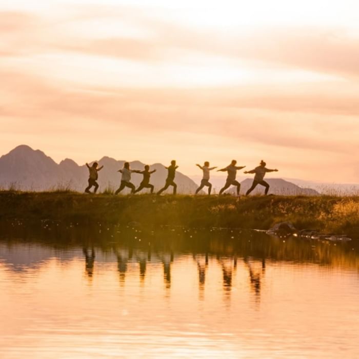 Gruppe praktiziert Yoga am Seeufer bei Sonnenuntergang mit Bergkulisse im Hintergrund bei Falkensteiner Hotel & Spa Carinzia