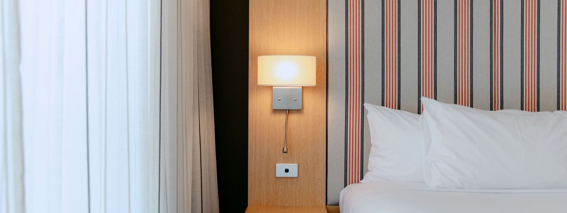 Hotel room with white bed, striped wall, white curtains, and a wall lamp with a switch.