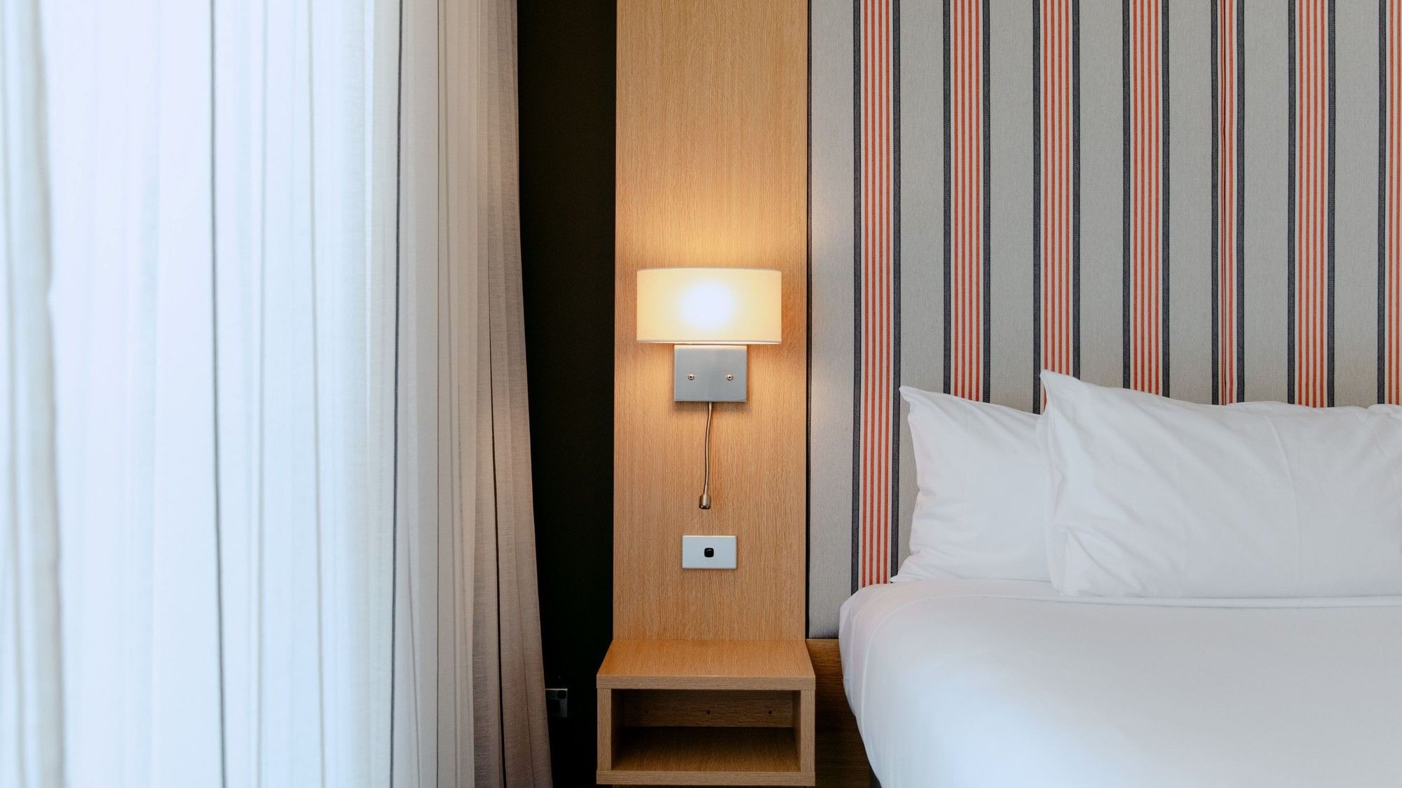 Hotel room with white bed, striped wall, white curtains, and a wall lamp with a switch.