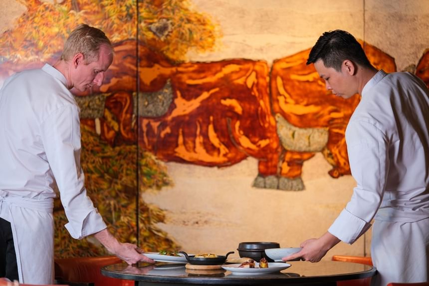 Two chefs placing gourmet dishes on a round table, set against a vibrant mural of a cow at Park Hyatt Saigon