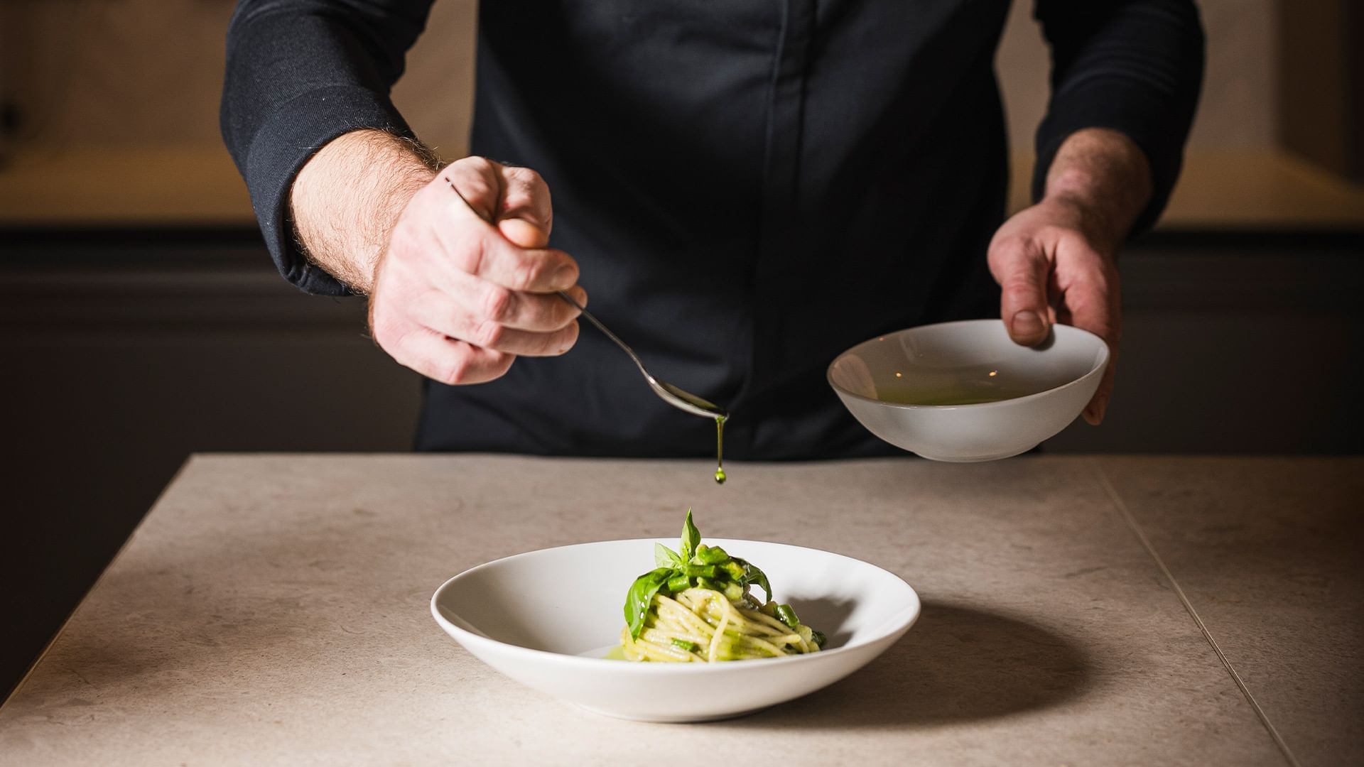 Chef in camicia nera versa condimento da una ciotola su una pasta verde in un'altra ciotola.
