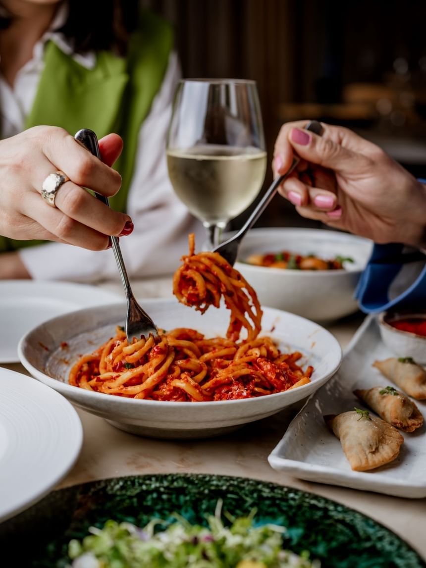Two hands twirl pasta from a bowl, accompanied by a glass of white wine on a table at The Kitchens