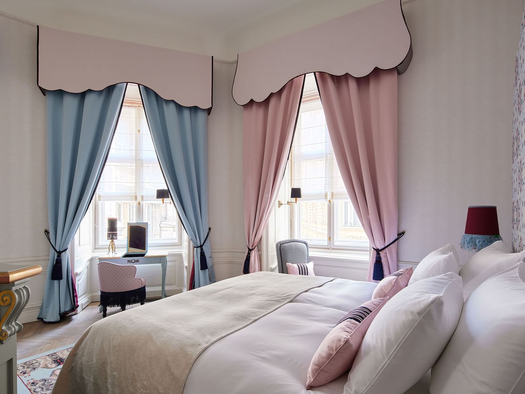 Elegant hotel bedroom with white bed, blue and pink curtains, floral wallpaper, and a small desk by the window.