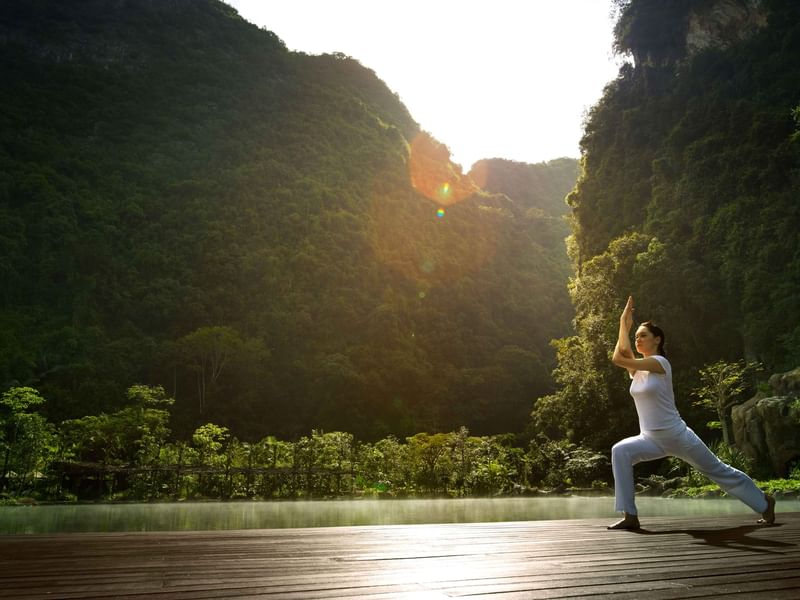 Woman practicing yoga on sky deck beside tranquil lake surrounded by lush green mountains at The Banjaran Hotsprings Retreat promoting Wellness Journey