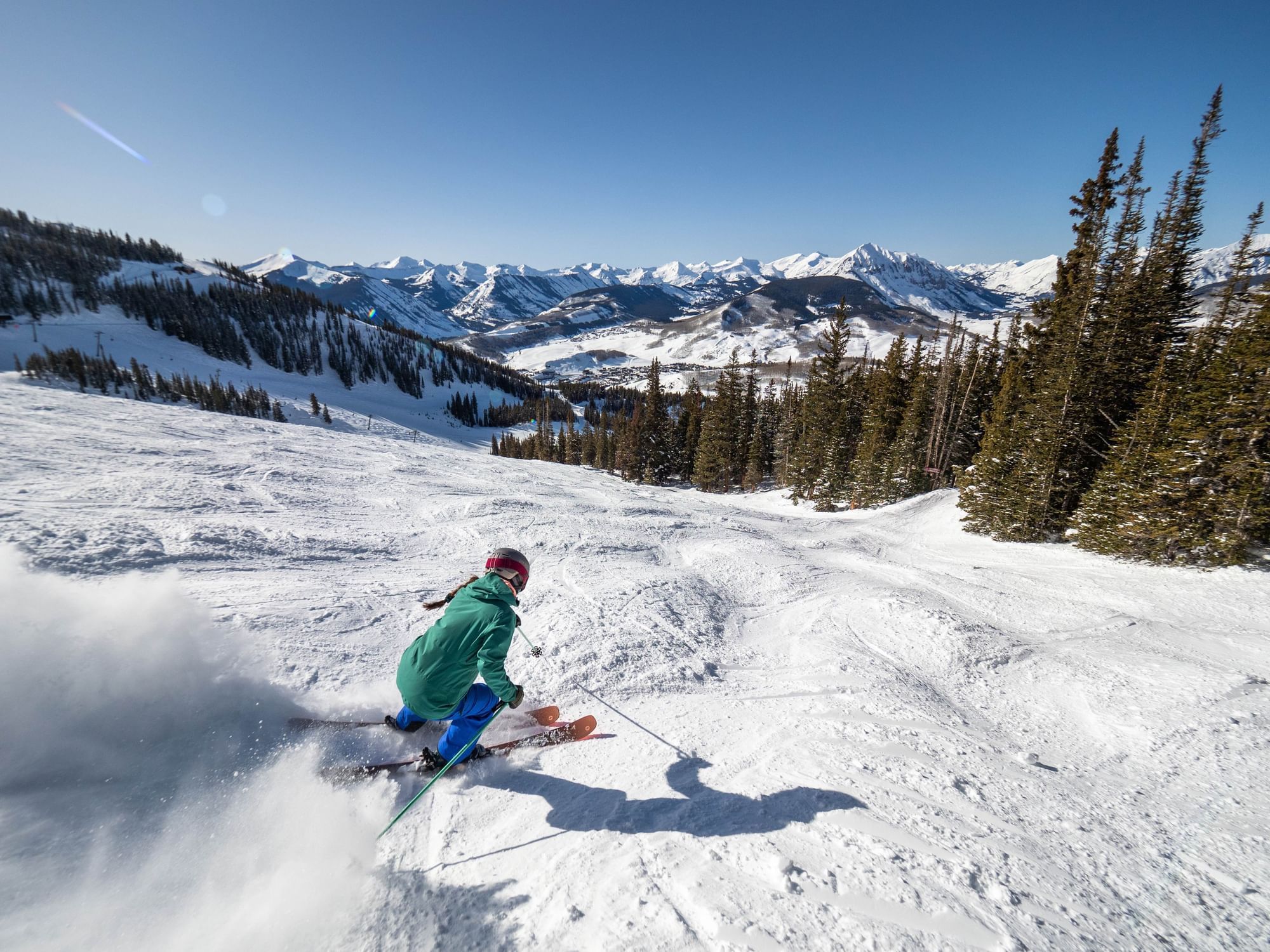 Skier in green jacket and blue pants skiing down slope with trees and mountains in background.