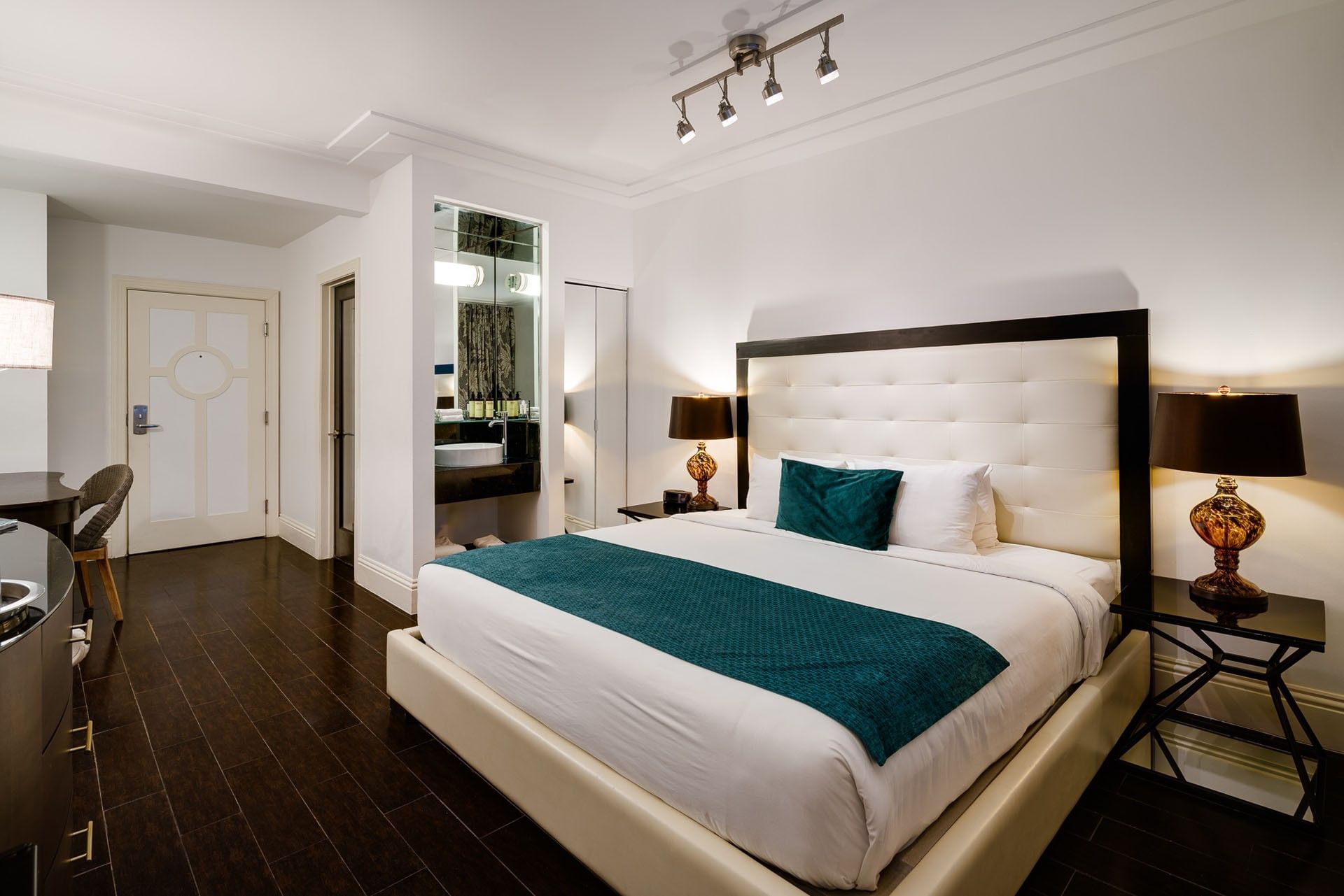 A large hotel room with a king-size bed, two nightstands with lamps, and a dark wood floor.