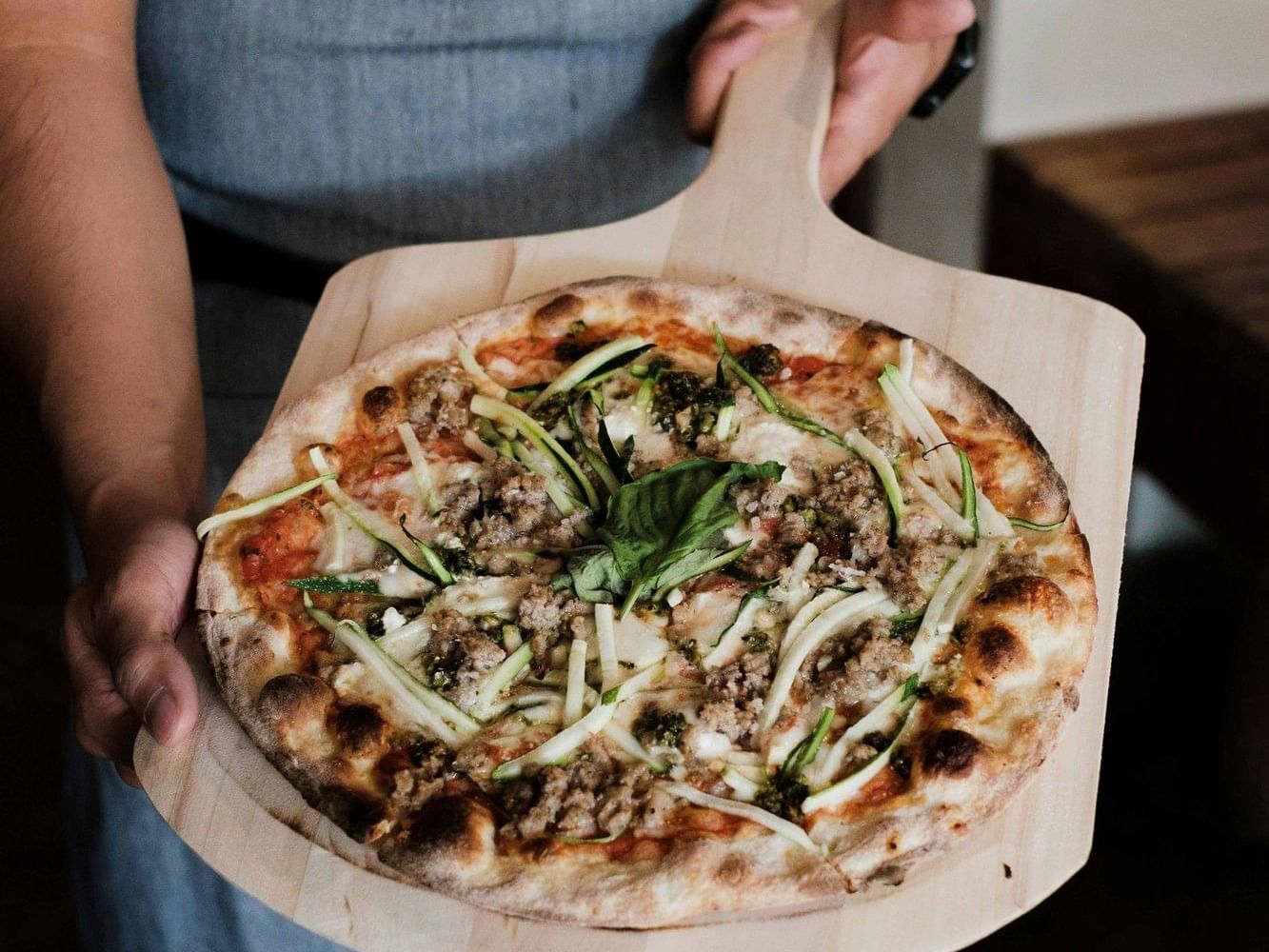 Staff member in an apron holding a freshly baked gourmet pizza on a wooden serving board, topped with herbs and cheese.