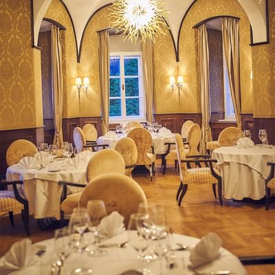 Elegantes Restaurant mit gedeckten Tischen, goldenen Stühlen, großen Fenstern und einer Kristallleuchter.