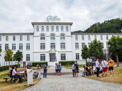 Exterior of Maison Cailler surrounded by lush greenery, and visitors near Hotel des Innovations