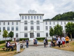 Exterior of Maison Cailler surrounded by lush greenery, and visitors near Hotel des Innovations