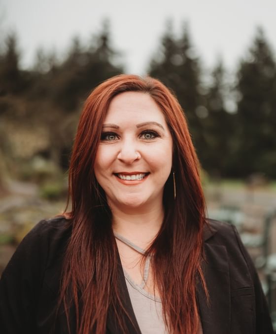 Gabby Palumbo smiling in a black blazer, standing by tall pine trees under an overcast sky at Oregon Garden Resort
