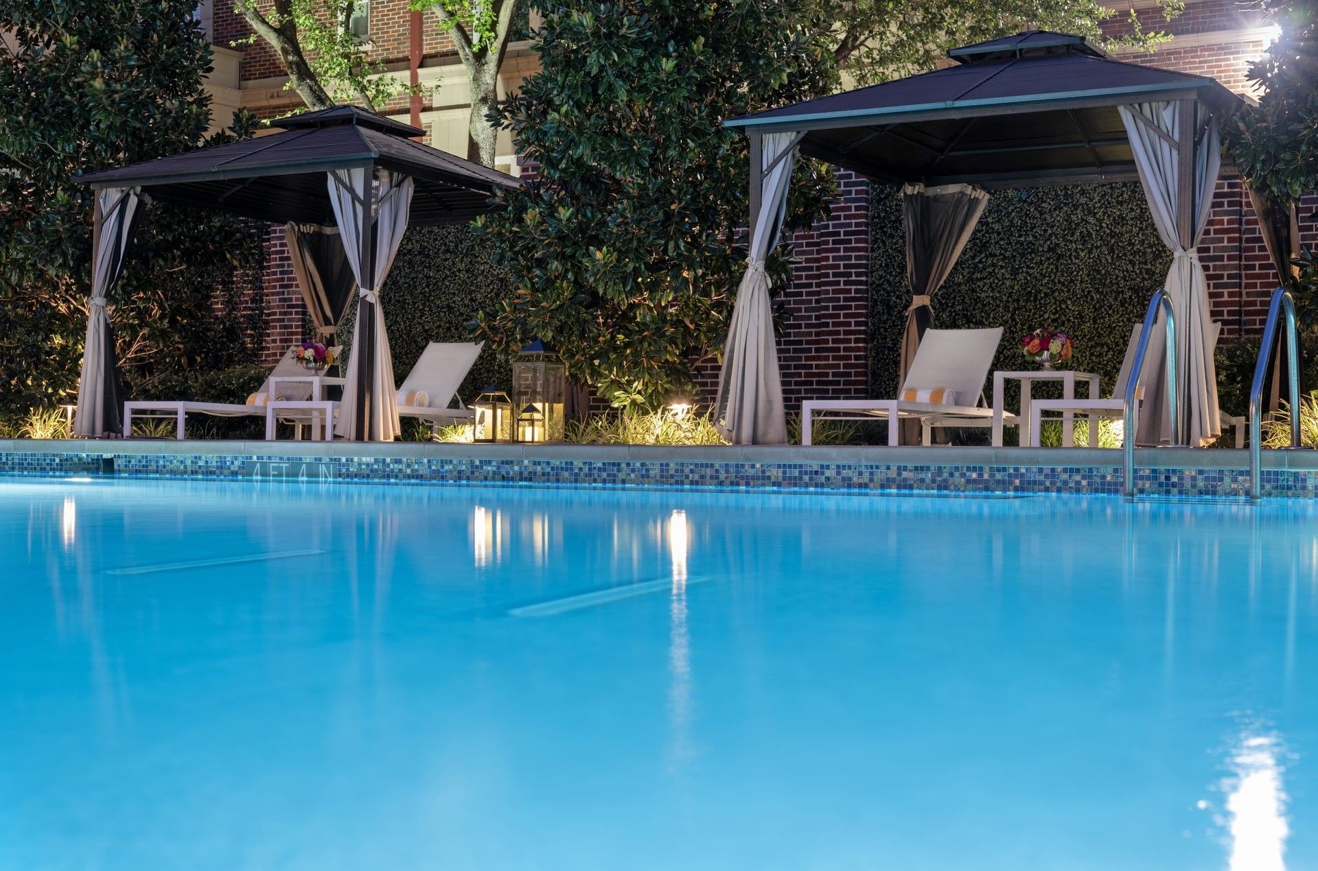 Lounge chairs by a table under a dark cabana surrounding the outdoor pool at Warwick Melrose Dallas