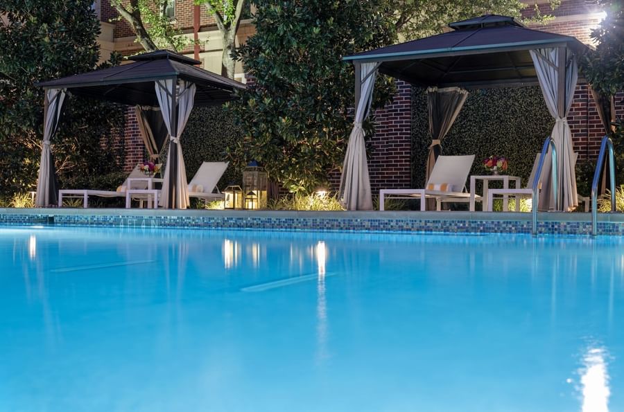 Lounge chairs by a table under a dark cabana surrounding the outdoor pool at Warwick Melrose Dallas