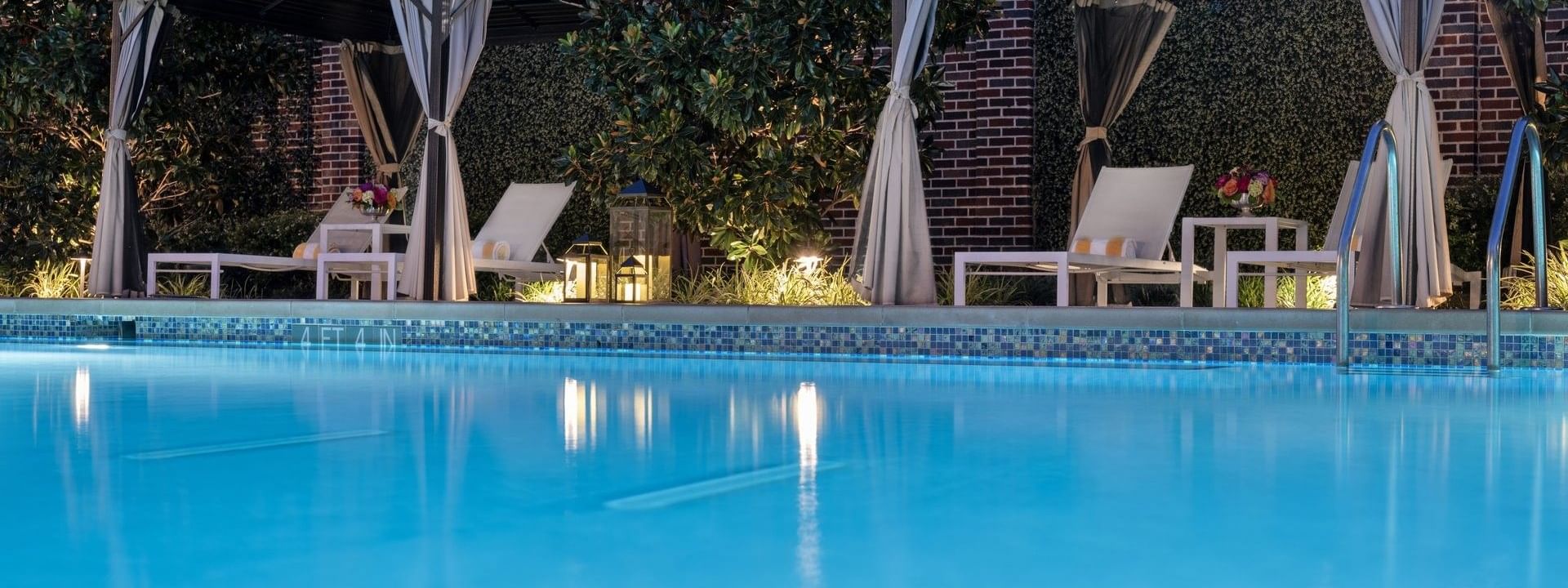 Blue pool by lounge chairs under cabanas surrounding the lit courtyard garden at Warwick Melrose Dallas
