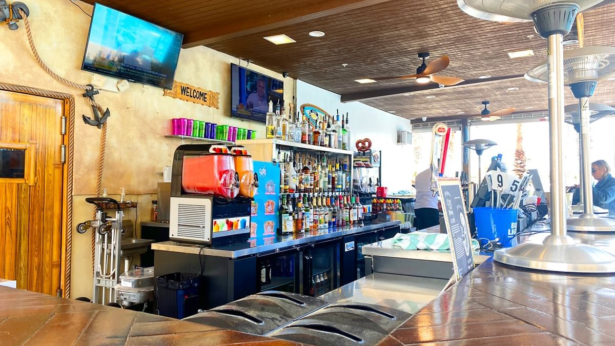 A bar with a counter, several bottles, a tv, a heater, and a ceiling fan.