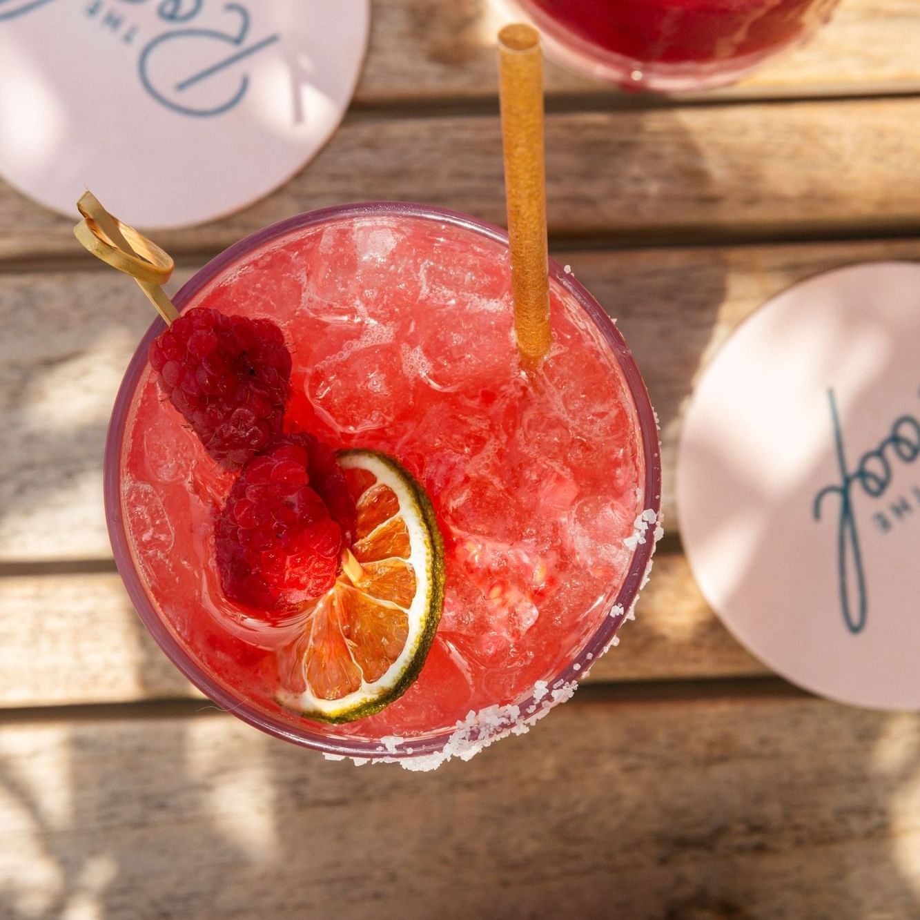 Two colorful cocktails with fruit garnishes and straws on a wooden table with branded coasters.