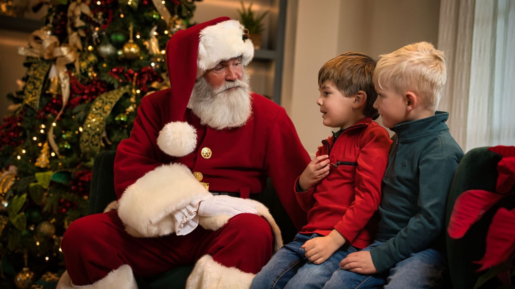 Santa Claus sits with two young boys, one in red and one in green, near a decorated Christmas tree.