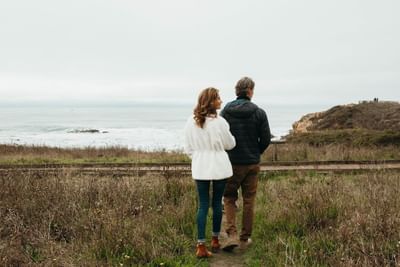 A couple walks along a coastal trail, looking out at the ocean on a cloudy day.