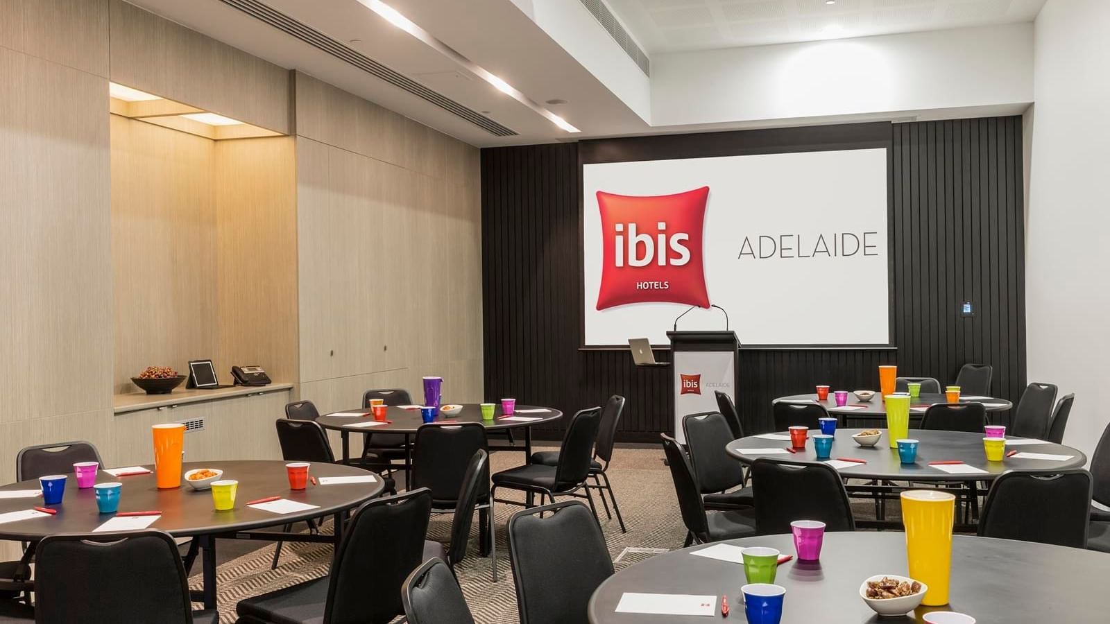 Round tables set up with chairs facing a large projection screen in The Think Tank at Ibis Adelaide