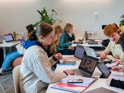 Students collaborating at white tables with laptops and notebooks in a bright modern study space with green plants.