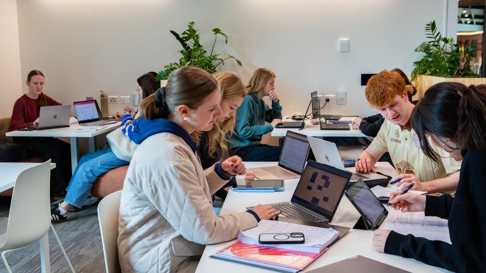 Students collaborating at white tables with laptops and notebooks in a bright modern study space with green plants.