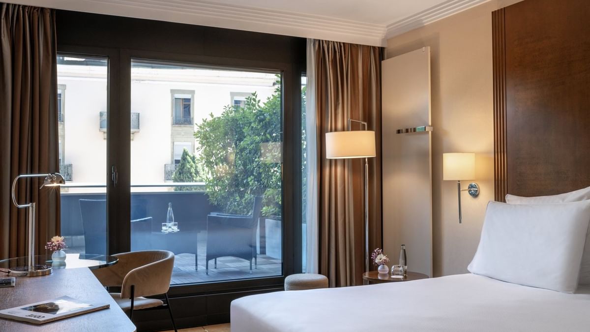 Plush bed by a wooden desk with a lamp, facing a terrace in Deluxe Terrace Room at Warwick Geneva