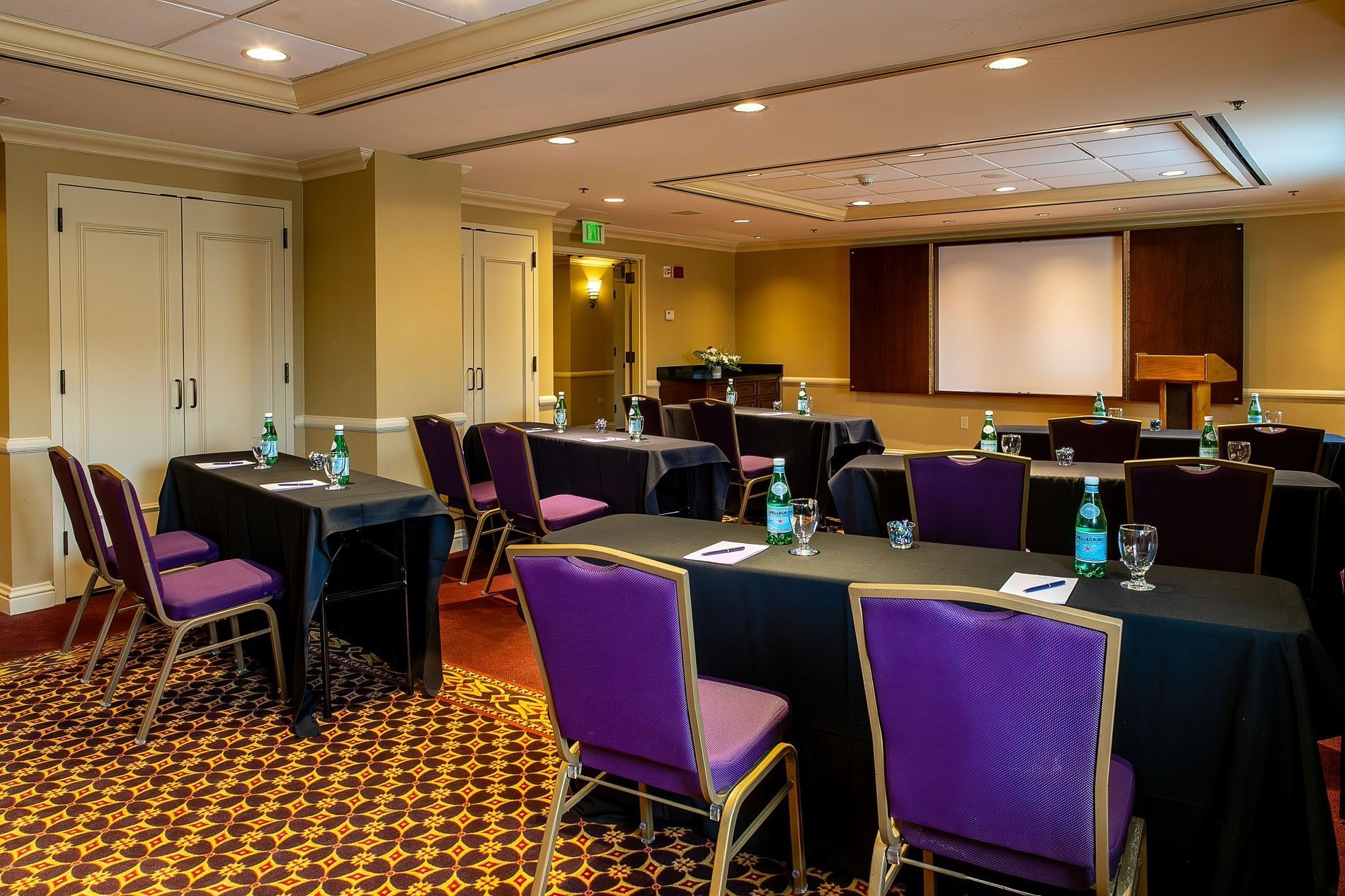 Purple chairs by black tables under a projector screen near a wooden lectern in Tabor II at Warwick Denver